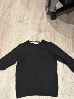 Svart stickad tröja från J.Lindeberg - Snygg svart stickad tröja från J.Lindeberg med diskret logga på bröstet. Tröjan har rund halsringning, långa ärmar och ribbade muddar vid ärmslut och nederkant. Perfekt basplagg till garderoben.