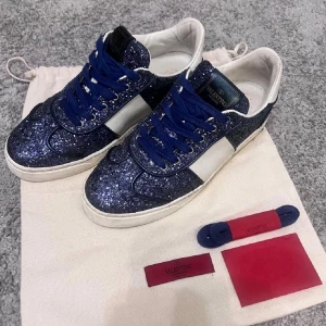 Valentino blå glittriga sneakers toppskick  - Snygga sneakers från Valentino med blå glitterdetaljer, vita paneler och blå skosnören. Skorna har en klassisk låg modell och en vit sula. Märkeslogga på plösen och extra skosnören medföljer. Perfekta för dig som vill sticka ut med stil. 10/10 unisex sneakers storlek 38! Unik vara till en unik slant, passa på! Först t kvarn. 