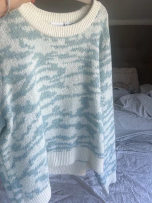 Ljusblå och vit mönstrad stickad tröja - Mysig stickad tröja i ljusblått och vitt med ett zebrainspirerat mönster. Tröjan har rund halsringning och ribbade muddar vid ärmslut och nederkant. Perfekt för dig som gillar en avslappnad och trendig stil.