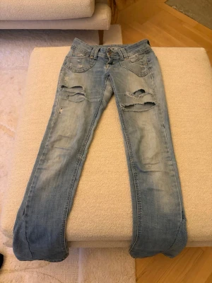 Ljusblå slitna jeans med dragkedjor - Säljer ett par ljusblå utsvängda jeans med slitningar och hål. Obs!! Kolla sista bilden för en defekt. 