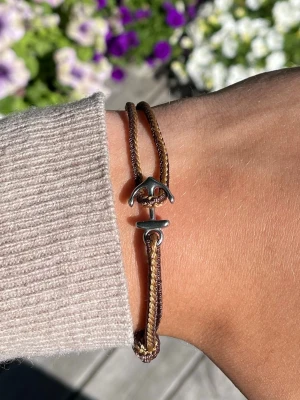 Brunt armband med ankardetalj - Snyggt armband i brunt flätat snöre med en silverfärgad ankardetalj som lås. Armbandet har en enkel och stilren design som passar perfekt till vardagslooken. Justerbar storlek tack vare knutarna. Perfekt accessoar för dig som gillar marina detaljer.