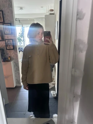 Beige fleece-tröja med halv dragkedja - Mysig beige fleece-tröja med halv dragkedja och hög krage. Tröjan har en avslappnad passform och ett litet broderat märke på bröstet. Perfekt för lager-på-lager och chill dagar. Materialet är mjukt och värmande fleece.