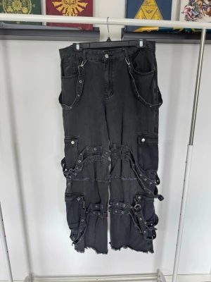 Svarta bondage jeans med straps - Unika svarta bondage jeans med flera straps, spännen och råa kanter. Tillverkade i denim med detaljer i metall.