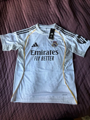 Real Madrid matchtröja Mbappé Adidas - Snygg vit Real Madrid matchtröja från Adidas med svarta detaljer och gula linjer. Tryck med Emirates Fly Better på framsidan och Mbappé 10 på ryggen. Officiell klubbmärke på bröstet och Adidas-logga. Perfekt för fotbollsfans och samlare.