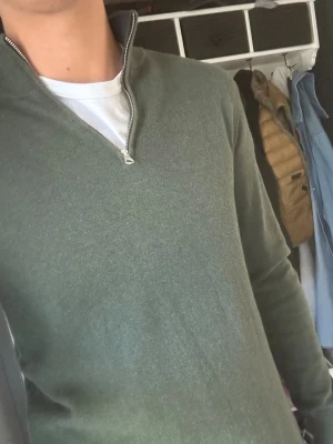John Henric half zip - Mörkgrön John Henric långärmad tröja med halv dragkedja i halsen. Tröjan har en enkel och stilren design med ribbade muddar vid ärmslut och nederkant. Perfekt att bära över en t-shirt för en avslappnad look. Materialet känns mjukt och bekvämt.