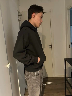 Svart Essentials hoodie Fear of God - Svart hoodie från Fear of God Essentials med stor logotyp på ryggen och mindre logotyp på bröstet. Klassisk passform med huva och ribbade muddar. Materialet är mjukt och bekvämt, perfekt för en avslappnad look.