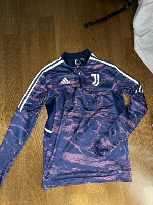 Juventus träningströja från Adidas - Juventus långärmad träningströja från Adidas i marinblått med rosa mönster och vita detaljer. Tröjan har halv dragkedja, klubbmärke på bröstet och klassiska Adidas-ränder på axlarna. Tillverkad i funktionsmaterial som andas.