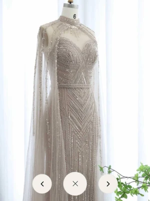 Champagnefärgad långklänning med pärlor - Elegant långklänning i champagnefärg med glittriga pärldetaljer och geometriskt mönster. Klänningen har hög hals, transparent cape och figurnära passform. Perfekt för bal eller fest. Materialet är troligtvis polyester eller syntet.