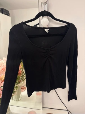 Svart långärmad topp med rynk - Säljer en svart långärmad topp med snygg rynkdetalj framtill och djupare ringning. Modellen är tight och passar perfekt till både jeans och kjol. Materialet känns mjukt och stretchigt, troligen bomull eller polyester.