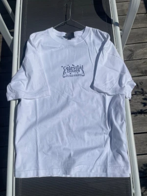 Vit t-shirt - Säljer en vit t-shirt med tryck både fram och bak. Framsidan har en liten logga och baksidan har ett stort grafiskt tryck med planetmotiv och text. Klassisk passform och rund hals. Perfekt för dig som gillar unika prints och streetwear vibes.
