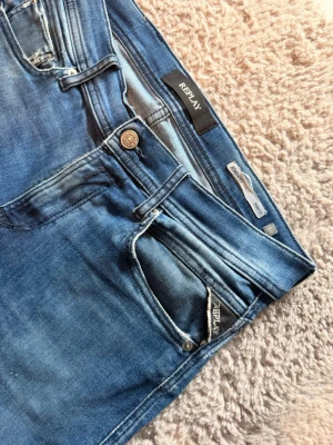 Blå slim fit jeans från Replay - Snygga blå slim fit jeans från Replay med klassisk femficksmodell och slitna detaljer. Jeansen har en något tvättad look och smala ben. Tillverkade i stretchigt denim för extra komfort. Perfekta för en trendig och avslappnad stil.                             Det finns små slitage men enligt min och många andras åsikter så blir replay jeans bara snyggare så🤩🤩 Skriv vid minsta lilla fundering✅