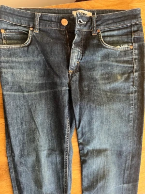 Blå jeans från Acne Studios - Snygga blå jeans från Acne Studios med klassisk femficksmodell och dragkedjegylf. Jeansen har en tvättad look med lätt slitning vid fickan och kopparfärgade nitar. Perfekta för en avslappnad och trendig stil.