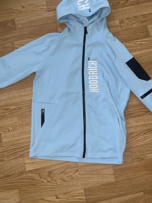 Ljusblå hoodie från Hoodrich - Snygg ljusblå hoodie från Hoodrich med dragkedja och huva. Stora vita tryck på bröstet och ryggen, samt svart detalj på ena ärmen. Två fickor med dragkedja framtill. Perfekt för en streetwear-look. Byxorna är helt nya men hoodien är använd max 2 gånger. Priset går och diskuteras vid snabb affär.