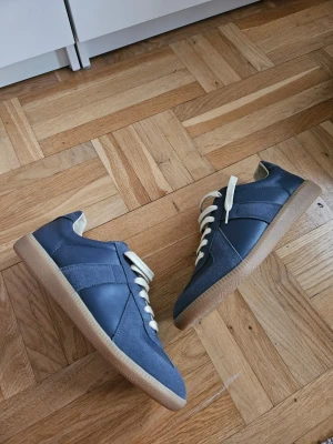 Maison Margiela Pewter Gat - Snygga Maison Margiela mörkblå sneakers med ovandel i både mocka och läder, beigea skosnören och klassisk gummisula. Skorna har en stilren design med paneler i olika material och passar perfekt till streetwear eller casual outfits.