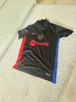 FC Barcelona svart matchtröja Nike - Snygg svart FC Barcelona matchtröja från Nike med röda och blå detaljer på sidorna. Tröjan har klubbmärke, Nike-logga och Spotify-tryck på bröstet samt UNHCR-logga på ryggen. Tillverkad i lätt och ventilerande polyester, perfekt för fotbollsträning.