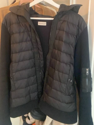 Svart pufferjacka från Moncler - Snygg svart pufferjacka från Moncler med quiltad front och huva. Jackan har ribbstickade ärmar med dragkedjeficka och Moncler-logga på armen. Perfekt för kyliga dagar och har dragkedja framtill. (Defekter-dragkedja är trasig men går att fixa billigt)