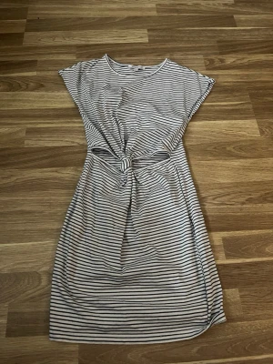 Randig knytklänning i bomull - Kortärmad klänning med smala svarta och vita ränder, knytning i midjan och rund halsringning. Avslappnad passform och mjukt bomullsmaterial, perfekt för en trendig och bekväm look.