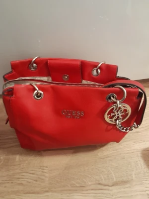 Röd handväska från Guess - Snygg röd handväska från Guess med silverfärgade metalldetaljer och logotyp framtill. Väskan har dragkedja upptill, dekorativ metallberlock och kedjedetalj på handtaget. Perfekt för dig som vill sticka ut med en färgstark accessoar.finns även matchande läderhandskar till
