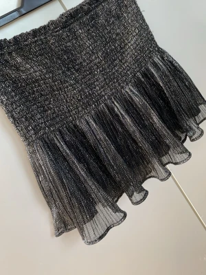 Glittrig svart kortkjol med volang - Supersnygg svart kortkjol med glittrigt tyg och volangkant. Kjolen har resår i midjan och ett skirt, lätt material som ger en festlig känsla. Perfekt för dig som vill sticka ut lite extra med metallic vibes och volanger.