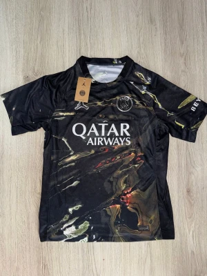 Svart PSG x Jordan matchtröja - Snygg svart PSG matchtröja i samarbete med Jordan, med unikt marmorerat mönster i gult och rött. Stort vitt Qatar Airways-tryck på bröstet och PSG-logga. Tillverkad i lätt och ventilerande funktionsmaterial, perfekt för träning eller match.