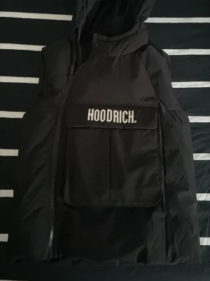 Svart dunväst från Hoodrich XL - Snygg svart dunväst från Hoodrich med stor ficka framtill och tydlig logga på locket. Västen har huva och dragkedja i sidan för en modern look. Perfekt för lager-på-lager och streetwear-stil. (Barnstorlek)