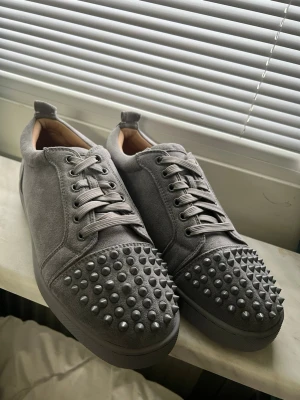 Gråa Louboutin sneakers  - Säljer ett par gråa Loubs i mocka med coola nitar på tån. Skorna har snörning och en låg siluett. Insidan är fodrad i beige läder för extra komfort. Perfekta för dig som vill sticka ut med dinn stil.