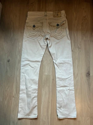 Vita jeans från True Religion - Säljer ett par vita jeans från True Religion med raka ben och klassiska kontrastsömmar. Jeansen har fem fickor, knappgylf och detaljerade bakfickor med knappar. Perfekta för dig som gillar streetwear och vill sticka ut lite extra.