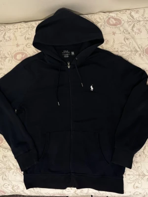 Navy full zip från Polo Ralph Lauren -  med dragkedja från Polo Ralph Lauren. Klassisk design med vit broderad logga på bröstet, huva med dragsko och kängurufickor framtill. Tillverkad i mjukt material, perfekt för en avslappnad och stilren look.