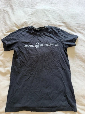 Svart Sail Racing t-shirt - Snygg svart t-shirt från Sail Racing med vit logga tryckt över bröstet. Klassisk rund halsringning och korta ärmar. Tillverkad i mjuk bomull som känns skön mot huden. Perfekt för en avslappnad och sportig stil.