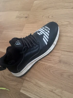 Svarta sneakers från Emporio Armani - Snygga svarta sneakers från Emporio Armani med vit logga och GA-vinge på tån. Skorna har svarta snören, vit sula med svart undersida och är tillverkade i syntetmaterial och textil. Perfekta för dig som vill ha en stilren och modern look även finskor