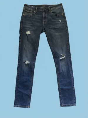 Mörkblå jeans med slitningar. - Säljer ett par mörkblå jeans med raka ben och flera slitningar framtill för en trendig look. Jeansen har klassisk femficksdesign och knappgylf. Perfekta för dig som gillar en avslappnad och cool stil.