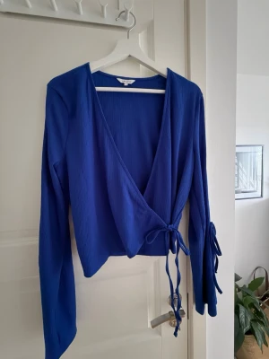 Blå omlottopp från Bubbleroom - Snygg blå omlottopp från Bubbleroom med långa ärmar och knyt i sidan. Toppen har ribbad struktur och v-ringad halsringning. Perfekt att styla med både jeans och kjol för en trendig look.