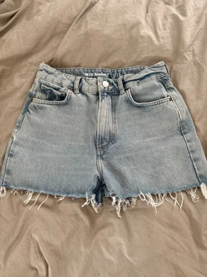 Blå jeansshorts med fransig kant - Säljer ett par klassiska blå jeansshorts i midwaist-modell med rå, fransig kant nertill. Shortsen har fem fickor, bälteshällor och stängs med knapp och dragkedja. 