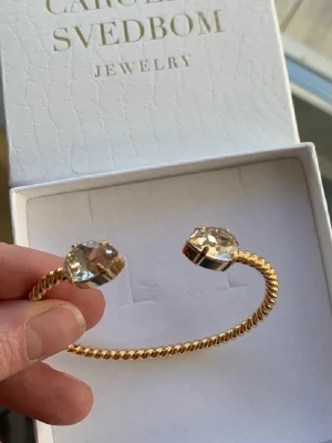 Guldarmband Caroline Svedbom - Snyggt armband från Caroline Svedbom i guldtonad metall med tvinnad design. Armbandet är öppet och pryds av två stora, gnistrande kristaller i båda ändar som ger en lyxig känsla. Perfekt accessoar för att lyfta din outfit.