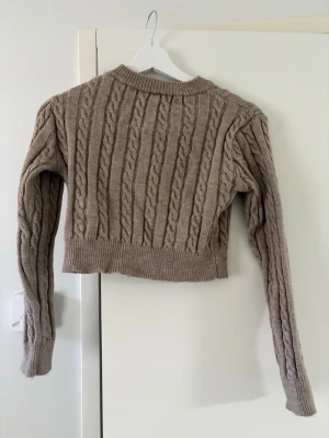 Beige kabelstickad cropped tröja - Säljer en beige kabelstickad tröja i croppad modell med långa ärmar och ribbade muddar. Tröjan har ett klassiskt flätmönster och är gjord i ett mjukt, tjockt material som känns riktigt cozy. Perfekt för lager på lager och snygg till både jeans och kjol.