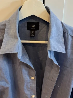 Blå skjorta från H&M med knappar - Klassisk blå skjorta från H&M i bomull med normal passform. Skjortan har långa ärmar, vit knappstängning framtill och en stilren krage. 
