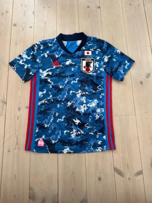 Japan blå kamouflage fotbollströja Adidas - Säljer en blå kamouflage fotbollströja från Adidas med Japans landslagsmärke på bröstet. Tröjan har röda och blå ränder på sidorna, mörkblå V-ringad krage och är tillverkad i ett lätt, ventilerande material. Perfekt för träning eller match!