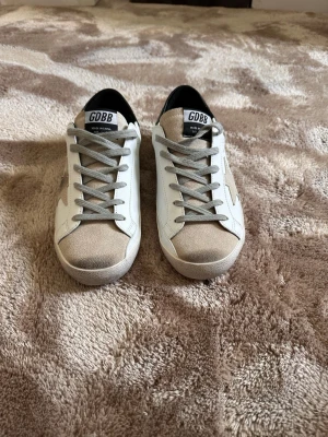 Golden goose - Snygga vita sneakers från GDBB med beige mockadetaljer på tån och en beige stjärna på sidan. Skorna har grå skosnören och svart hälparti. Tillverkade i en mix av läder och mocka för en trendig look.