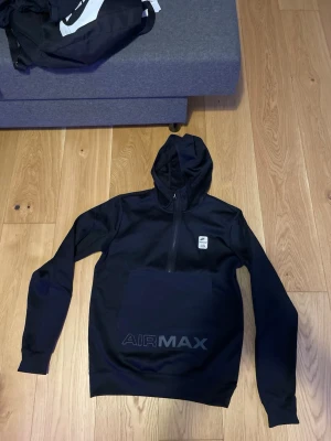 Svart Nike Air Max hoodie med huva - Snygg svart hoodie från Nike Air Max med huva och halv dragkedja framtill. Stor magficka med AIR MAX-tryck och liten Nike-logga på bröstet. Perfekt för en sportig och avslappnad stil.