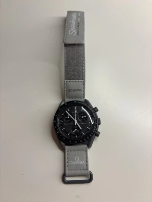 Omega x Swatch Speedmaster klocka - Stilren Omega x Swatch Speedmaster klocka med svart urtavla, kronograf och grått band. Klockan har tydliga vita markeringar och detaljer samt loggor på bandet. Baksidan har en måndekor och text. Perfekt för dig som gillar ikoniska samarbeten.