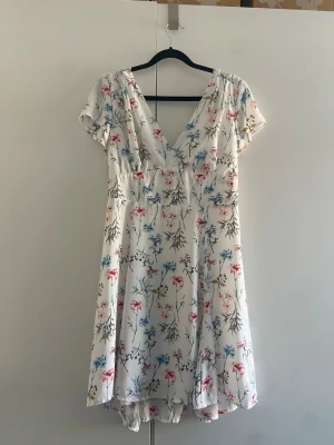 Vit blommig kortklänning från Bikbok  - Söt vit kortklänning med blommigt mönster i rosa, blått och grönt. Klänningen har v-ringning fram, korta ärmar och ett lätt, luftigt fall. Perfekt för varma dagar och ger en romantisk vibe.