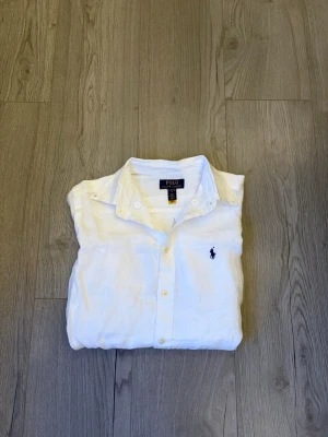 Vit linneskjorta från Polo Ralph Lauren - Klassisk vit skjorta i linne från Polo Ralph Lauren med blå broderad logga på bröstet. Skjortan har knappar framtill, lång ärm och en avslappnad passform. Perfekt till sommaren för en clean och stilren look. Den är helt ny! Det är storlek L i barnstorlek men jag skulle säga att den passar XS! Skriv vid minsta fundering!✅