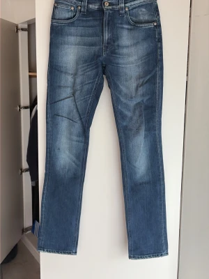 Blå raka / slimfit jeans från Nudie Jeans - Säljer ett par klassiska blå jeans från Nudie Jeans med raka / slimfit ben och snygg slitning. Jeansen har fem fickor och den ikoniska vågade sömmen på bakfickan. Tillverkade i mjuk denim med en skön passform, perfekta för en avslappnad stil.