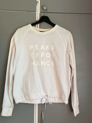Vit sweatshirt från Peak Performance - Vit sweatshirt med rund hals och tryckt logga 'PEAK PERFORMANCE' framtill. Tröjan har långa ärmar, ribbade muddar och dragsko i nederkanten för en snygg passform. Perfekt för en avslappnad och sportig stil.