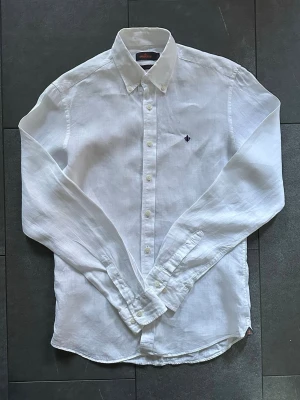 Vit linneskjorta från Morris - Klassisk vit skjorta från Morris i 100% linne. Skjortan har lång ärm, knappar framtill och en liten broderad logga på bröstet. Perfekt för en clean och stilren look, passar till både jeans och chinos.