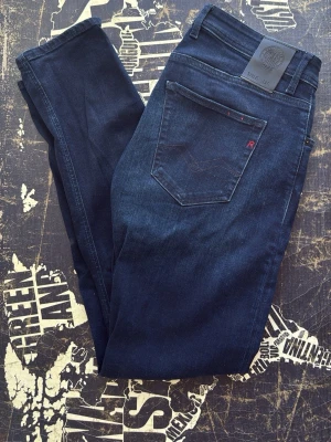 Mörkblå skinny jeans från Replay - Snygga mörkblå skinny jeans från Replay med klassisk femficksdesign och diskret röd detalj på bakfickan. Jeansen har en smal passform och är tillverkade i stretchigt denimtyg för extra komfort. Perfekta för en trendig och avslappnad stil.