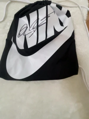 Signerad gympa påse av dejan kulusevski  - Snygg svart och vit gympåse från Nike. Signerad av Dejan kulusevski med stor logga framtill och dragskostängning. Tillverkad i lätt och slitstarkt material, perfekt för att bära träningskläder eller småprylar. Klassisk sportig design med vita snören.