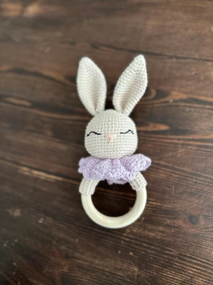 Virkad kanin skallra med lila krage - Söt handvirkad skallra i form av en kanin med stora öron och broderat ansikte. Kaninen har en ljuslila virkad krage och sitter fast på en rund träring. Perfekt som present till små barn eller som dekoration.