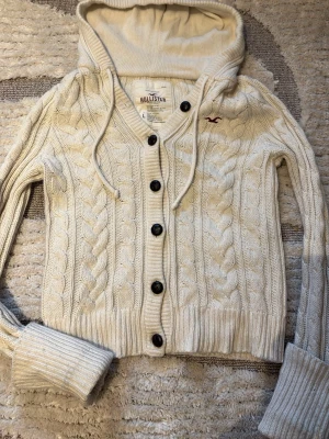 Hollister stickad hoodie🫶🏼 - Säljer en vit kabelstickad hoodie från Hollister med stora svarta knappar och dragsko i huvan. Tröjan har ribbade muddar, V-ringning och ett litet broderat logomärke på bröstet. Perfekt för dig som gillar mysiga och stilrena plagg. Det står att det är storlek L men den är liten så jag skulle säga S, ärmarna är däremot ganska långa så man kan vika upp de sådär som på bilden.