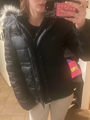 Hollister jacka - Säljer en  blå pufferjacka från Hollister  med stor huva och grå fuskpäls. Jackan har dragkedja och knappar framtill, två fickor och quiltad design. Perfekt för kalla vinterdagar och riktigt mysig att bära. 